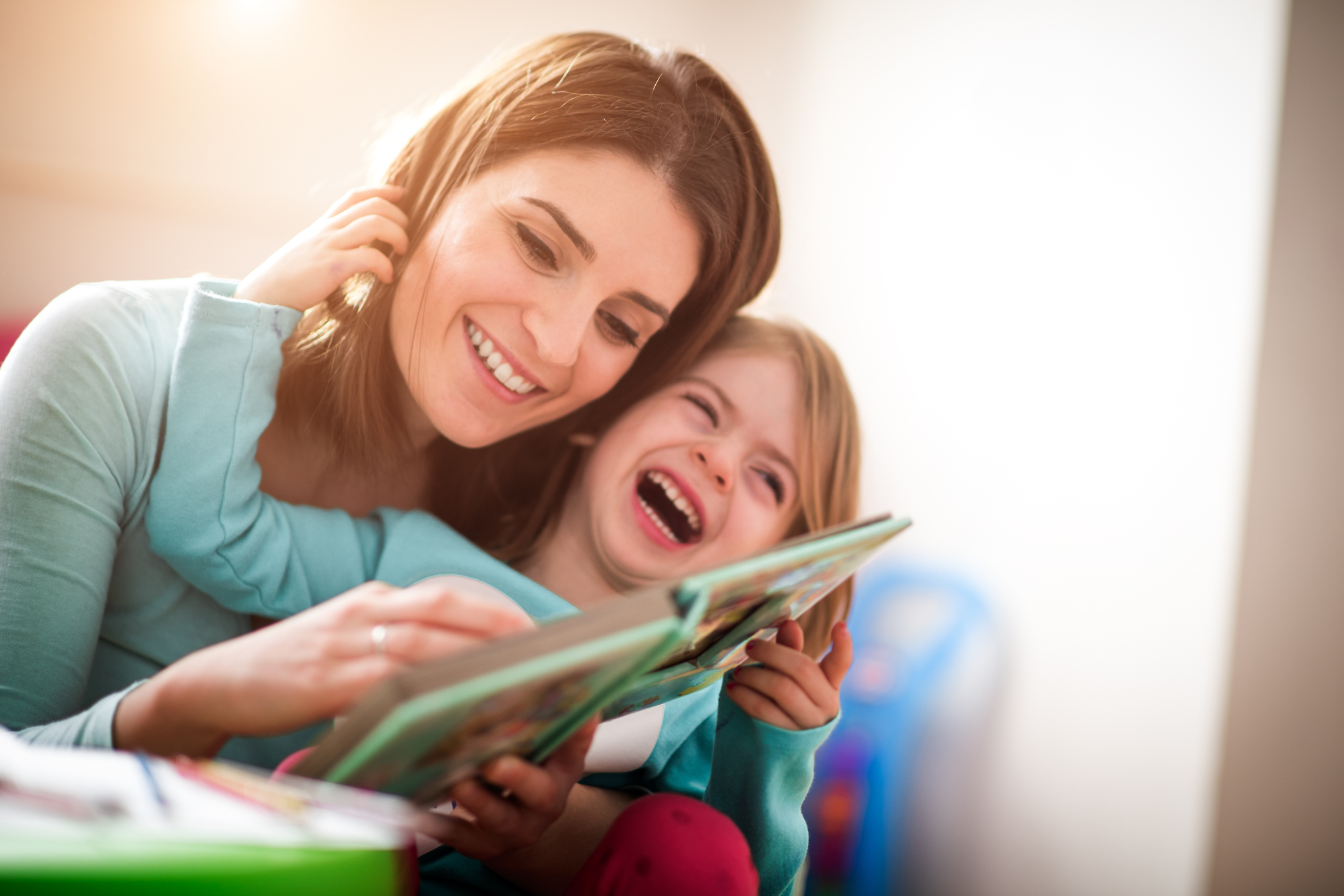 Mother and baby daughter at home reading a book and laughing together.-kidhours